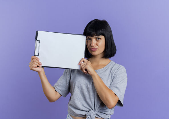 恼怒恼怒的年轻黑发白种女孩拿着剪贴板孤立在紫色背景与复制空间紫色空间剪贴板