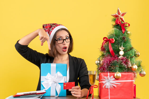 漂亮迷人的女士穿着西装 戴着圣诞老人的帽子和眼镜 在办公室里指着礼物和银行卡圣诞老人人圣诞老人