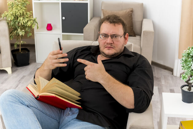腿自信的成年斯拉夫人戴着眼镜坐在扶手椅上 腿上拿着书 指着客厅里的电话坐着斯拉夫人眼镜