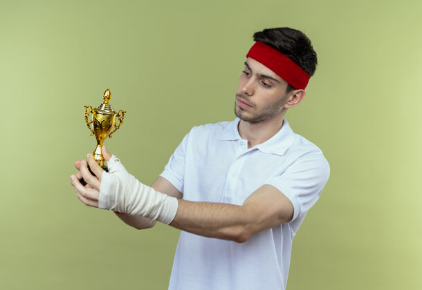 运动戴着头巾的年轻运动型男子拿着他的奖杯 站在绿色的背景下自信地看着奖杯持球站立年轻