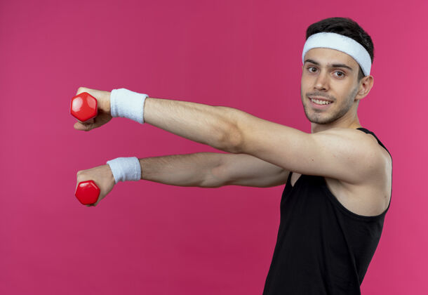 运动年轻的运动型男子戴着头巾 手持哑铃 站在粉色背景上 微笑着 快乐而积极工作头带微笑