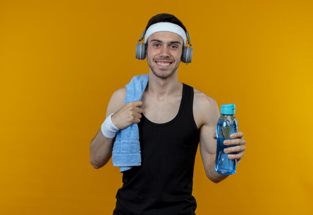 相机戴着头巾 肩上扛着毛巾 拿着一瓶水 站在橙色背景下微笑着看着摄像机的年轻运动型男子毛巾运动站着