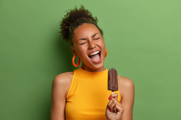 零食喜出望外的非洲裔美国妇女喜欢冰冻巧克力冰淇淋 张大嘴巴 在炎热的夏天玩得很开心 吃着美味的甜点 隔离在绿色的墙壁上垃圾食品 卡路里美味室内饥饿