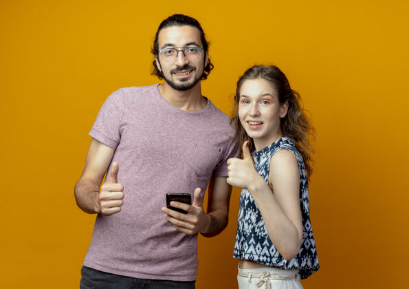 相机这对年轻夫妇看着镜头 微笑着 积极地竖起大拇指站在橙色的背景上站起来情侣拇指