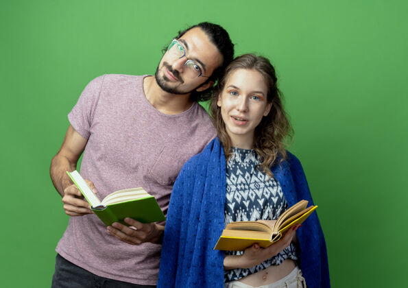 年轻人一对年轻夫妇拿着毯子 拿着书 站在绿色的背景上微笑着看着相机抱着站着微笑