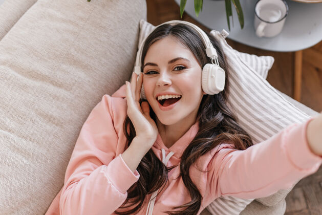漂亮穿着粉色运动衫的美女躺在沙发上 戴着耳机欣赏音乐 自拍抱着休闲人