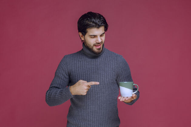 年轻一个穿着灰色毛衣的男人拿着一个咖啡杯指着它年轻人工人成人