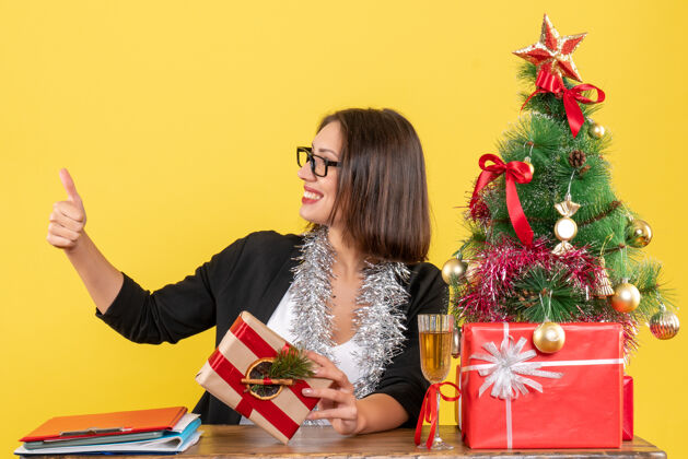 手势在办公室里 一位穿着西装戴着眼镜的漂亮商务女士正坐在一张桌子旁 桌上放着一棵圣诞树购物人微笑