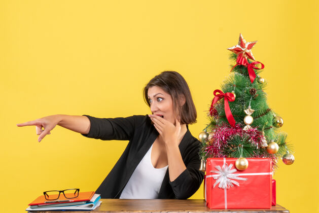 笔记本电脑一位年轻的女士坐在一张桌子旁 指着黄色办公室里装饰好的圣诞树旁西装革履的东西套装圣诞节坐