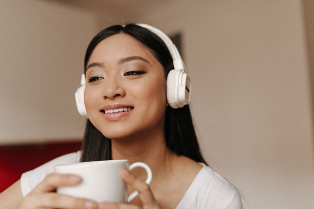 内部戴着大耳机的可爱亚洲女人微笑着端着一杯茶现代桌子空气