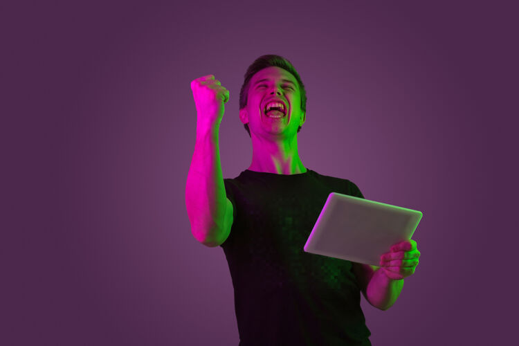 公司玩平板电脑 电子游戏 赢家霓虹灯下紫色工作室背景上的白种人肖像穿着黑色衬衫的漂亮男模人类情感的概念 面部表情 销售 广告面部肖像高管