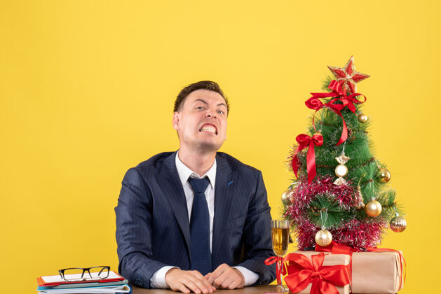 礼物前视图愤怒的男人坐在圣诞树旁边的桌子上 呈现黄色背景愤怒的男人会议前面