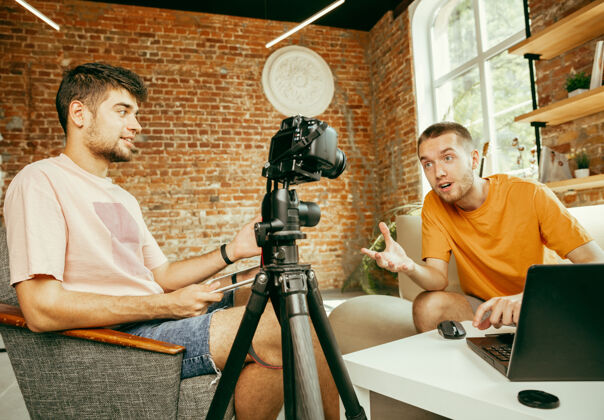 相机两位年轻的白人男性博主穿着休闲服 在家里用专业设备或摄像机录制视频采访博客 视频博客 视频博客在室内直播时聊天年轻博客室内