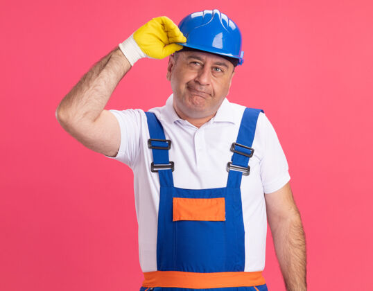 衣服体贴的成年建筑工人穿着制服 戴着防护手套 把安全帽隔离在粉红色的墙上安全穿着人