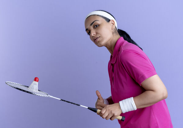 头带年轻自信的白人运动女性戴着头带和腕带站在一边拿着羽毛球在球拍上紫色背景与复制空间隔离穿自信立场