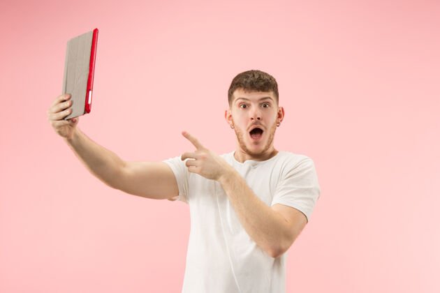 通信一张微笑的男人指着笔记本电脑的肖像 粉色工作室背景上的空白屏幕人类情感 面部表情概念和广告概念年轻肖像电脑