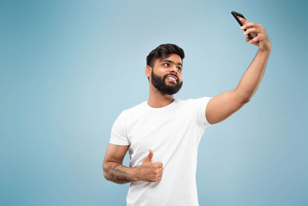 胡须蓝色背景上穿着白衬衫的印度教年轻人的半身特写肖像人类情感 面部表情 广告概念负面空间自拍或视频博客 视频博客 聊天选择企业家成功