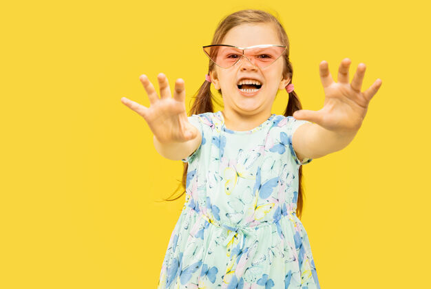 衣服美丽动情的小女孩孤立在黄色背景上半幅快乐的孩子站着的肖像 穿着裙子 戴着红色太阳镜夏天的概念 人类的情感 童年夏天长表情