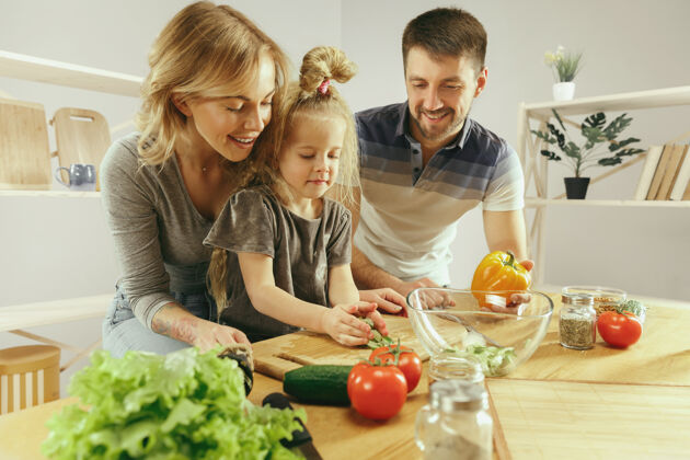一起可爱的小女孩和她美丽的父母正在家里的厨房里切菜 一边微笑着做沙拉家庭生活理念饭年轻午餐