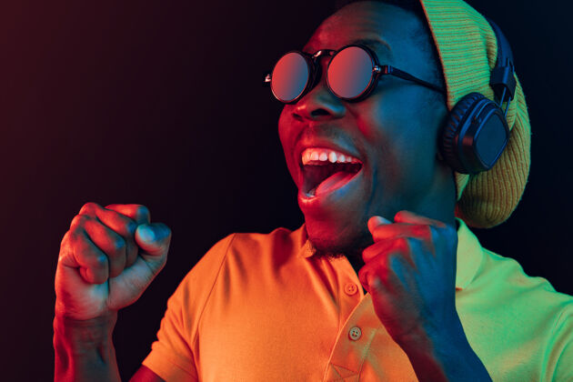 非洲年轻帅气快乐的时髦男人在霓虹灯下用耳机在黑工作室听音乐迪斯科舞厅 夜总会 嘻哈风格 积极的情绪 面部表情 舞蹈概念舞蹈男性家伙