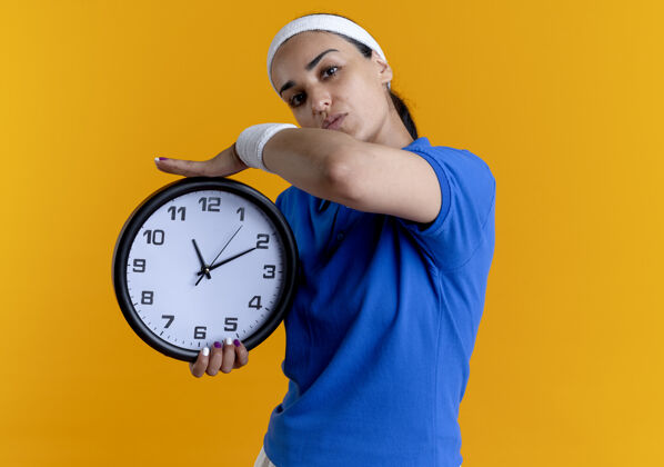 腕带年轻自信的白人运动女性戴着头带和腕带 拿着橙色背景上的时钟 并留有复制空间时钟自信手持