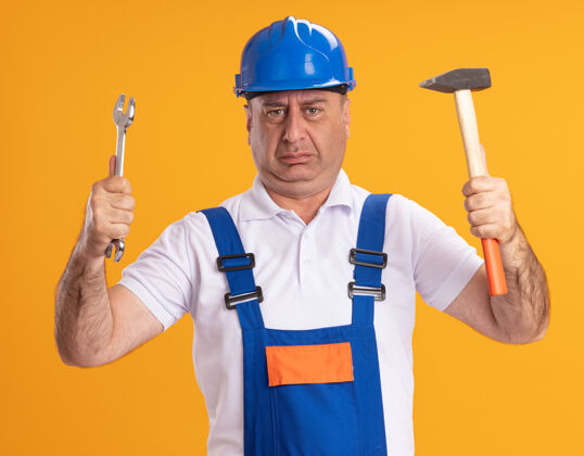 人穿着制服的未出租的成年建筑工人拿着隔离在橙色墙上的扳手和锤子成年人表情制服