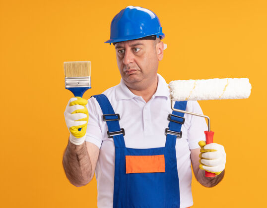 人困惑的成年建筑工人穿着制服 戴着防护手套 拿着油漆刷和滚筒刷隔离在橙色的墙上人人站立