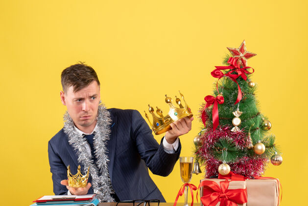 背景前视图商人比较他的皇冠坐在圣诞树附近的桌子上 黄色背景上的礼物生意人前面树