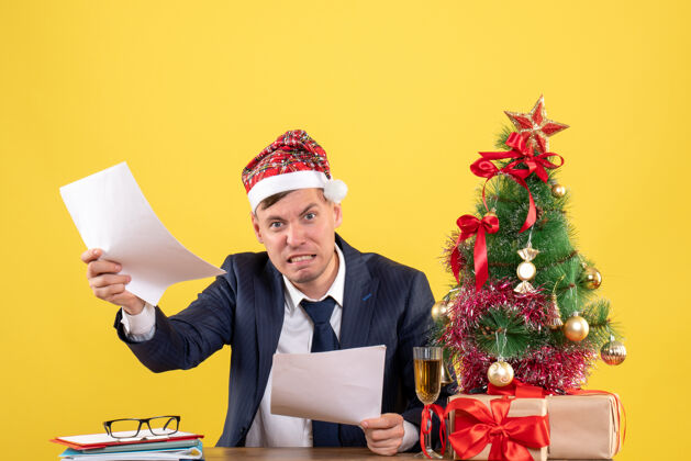 近前视图愤怒的人戴着圣诞帽坐在圣诞树旁的桌子旁 黄色背景上的礼物帽子圣诞老人行政人员