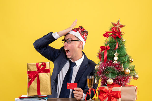 背景前视图惊奇的男人把手放在额头上坐在圣诞树旁的桌子上 呈现黄色背景树生意人