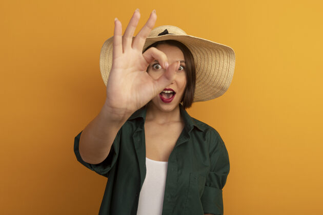 人兴奋的漂亮女人戴着沙滩帽 用手势表示同意 用手指隔着橘色的墙壁看前面通过手势帽子
