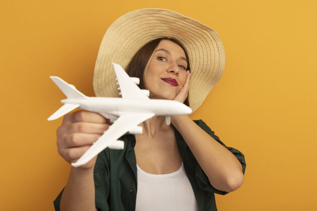 飞机戴着沙滩帽的漂亮女人把手放在脸上 把模型飞机孤立地放在橙色的墙上手请漂亮