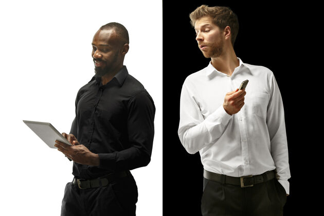 年轻成功快乐的黑人和白种人的男人与小玩意的混合夫妇在白色和黑色工作室的男模特的动态形象人类的面部情感概念关系笔记本电脑交流