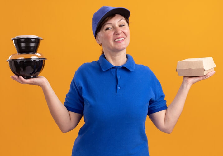 微笑身穿蓝色制服 头戴鸭舌帽的中年女送货员站在橘色的墙上 手里拿着食品包 开心而积极地看着前面帽子递送快乐