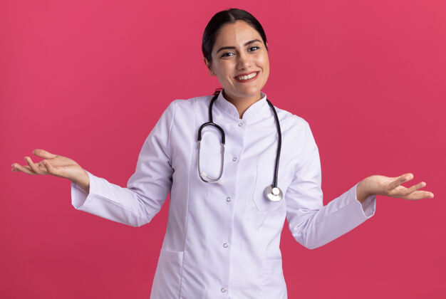 站立身穿医用外套 手持听诊器的年轻女医生站在粉红色的墙上 面带微笑 高举双臂 看着前方医生医生女人