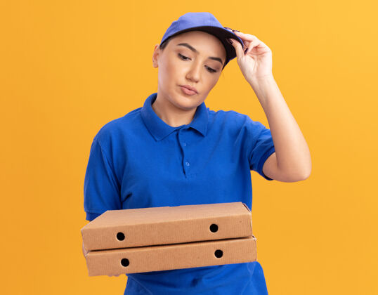 触摸身穿蓝色制服 头戴鸭舌帽的年轻女送货员拿着披萨盒 看着它们困惑地摸着站在橙色墙上的帽子盒子拿着女人