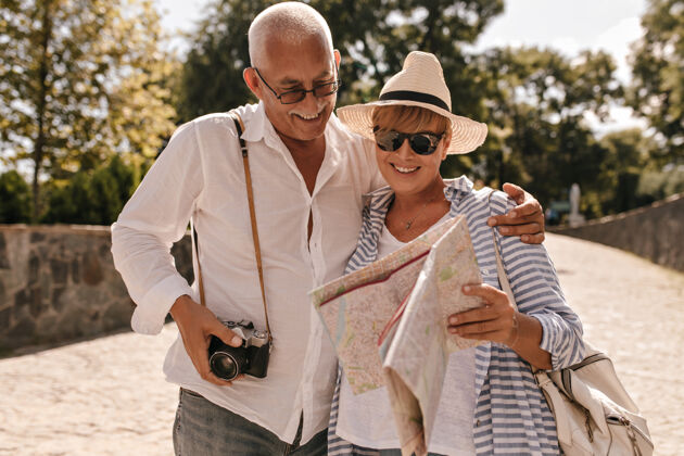 成年人一个快乐的男人 灰色头发 穿着浅色衬衫和牛仔裤 带着相机微笑 看着地图 金发女士戴着帽子 蓝色套装在公园里退休游客夫妇