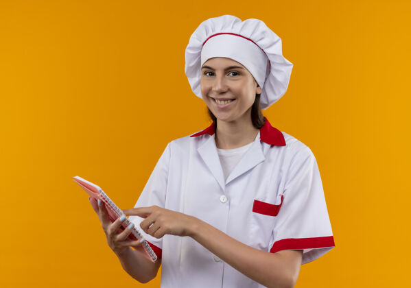 点身着厨师制服 面带微笑的白人年轻厨师女孩拿着并指着孤立在橙色背景上的笔记本 笔记本上有复印空间微笑笔记本厨师