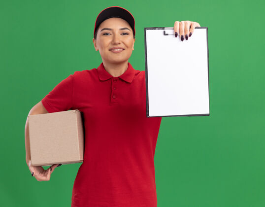 拿着身穿红色制服 头戴帽子的年轻女送货员拿着一个纸板箱 上面放着空白页的剪贴板 面带微笑地站在绿色的墙上看着前面欢呼年轻空白