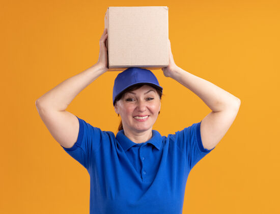 制服身穿蓝色制服 头戴鸭舌帽的中年女送货员举着纸板箱 站在橙色的墙上 面带微笑地看着前面抱着微笑欢呼