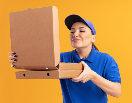 站着身着蓝色制服 戴着帽子的年轻送货员站在橘色的墙上 手里拿着披萨盒 打开盒子 吸入着怡人的香气拿着香气开着