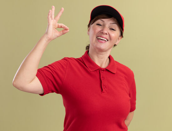 帽子身穿红色制服 头戴帽子的中年女送货员站在绿色的墙上 面带微笑 脸上挂着“ok”的牌子好中间年龄