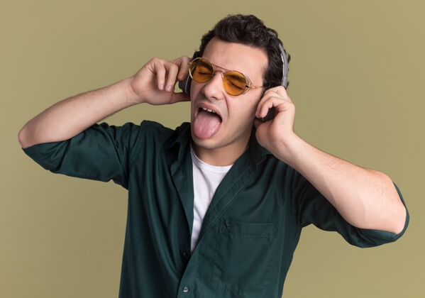 享受穿着绿色衬衫的快乐的年轻人戴着眼镜 戴着耳机 站在绿色的墙上欣赏着他最喜欢的音乐 伸出舌头眼镜穿男人