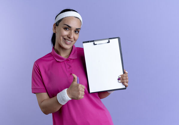拇指年轻微笑的白人运动女性戴着头带和腕带竖起大拇指拿着钢笔和剪贴板孤立的紫色背景与复制空间年轻穿剪贴板