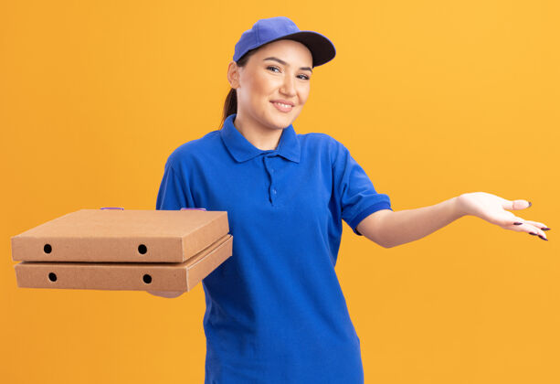 抱着身穿蓝色制服 头戴鸭舌帽 拿着比萨饼盒的年轻送货员面带微笑 站在橙色的墙上 一只手举着一张幸福的脸微笑脸手