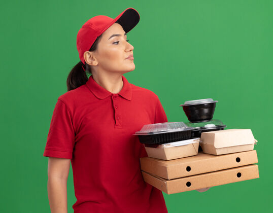 年轻人身穿红色制服 头戴鸭舌帽 拿着比萨饼盒和食品包的年轻女送货员站在绿色的墙上严肃地看着一旁女人严肃披萨