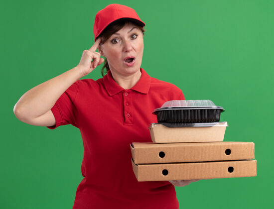 女人身穿红色制服 头戴鸭舌帽的中年女送货员拿着披萨盒和食品包 站在绿色的墙边困惑地看着前方中间立场制服