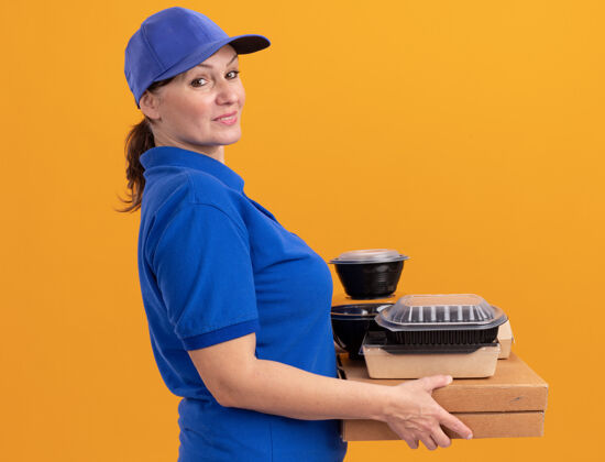 年龄身穿蓝色制服 头戴鸭舌帽的中年女送货员拿着披萨盒和食品包 微笑着自信地站在橙色的墙上看着前方披萨送货中年