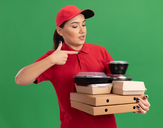 目录身穿红色制服 头戴鸭舌帽的年轻女送货员拿着披萨盒和食品包 用食指指着站在绿墙上困惑的她们女人手指站着