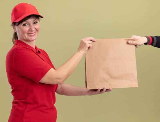 女人快乐的中年送货员身着红色制服 戴着帽子 站在绿色的墙边 友好地微笑着给顾客送纸包顾客中年帽子